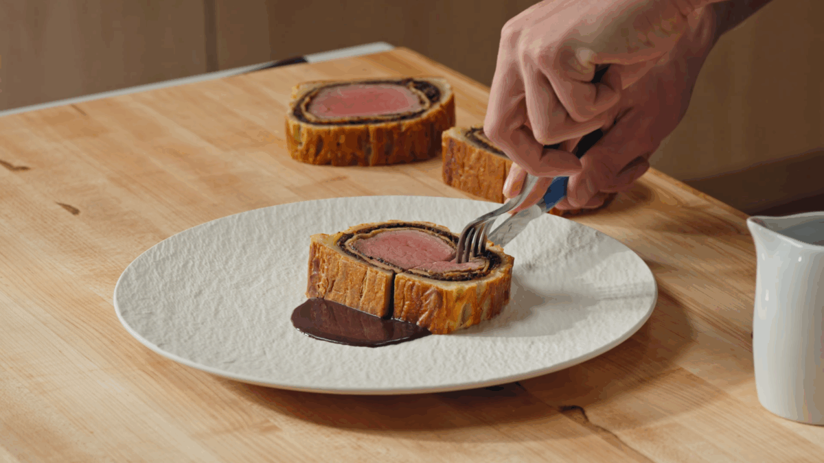 Nick DiGiovanni using a fork and knife to cut a bite off a slice of Beef Wellington on a plate. 
