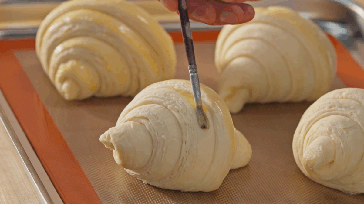 Nick DiGiovanni using a paint brush to brush egg wash on top of uncooked croissants.