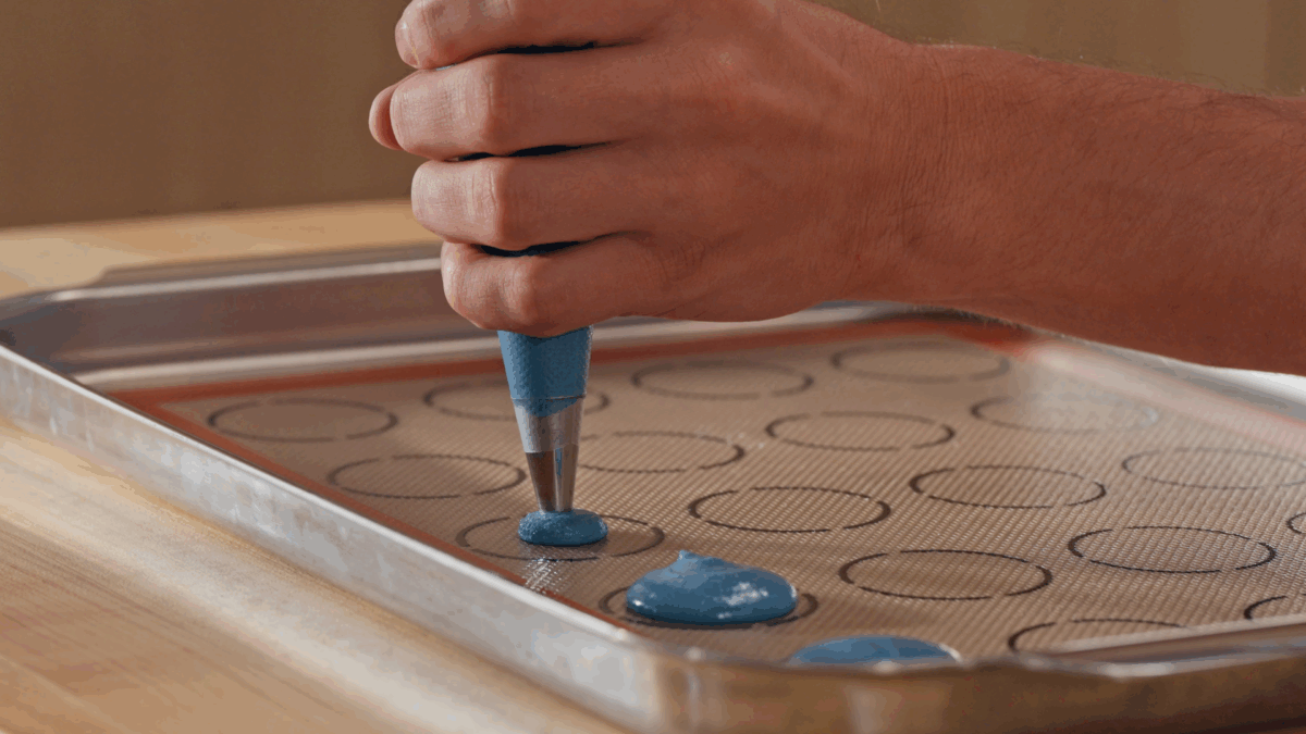 Nick DiGiovanni piping blue macaron batter onto a templated baking sheet. 
