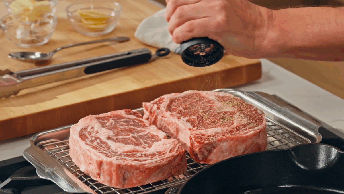 Nick DiGiovanni seasoning salted raw steaks with pepper.