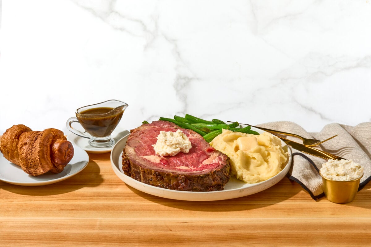 A slice of prime rib roast on a plate with mashed potatoes, and creamed spinach next to au jus and a Yorkshire pudding.