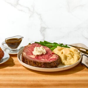 A slice of prime rib roast on a plate with mashed potatoes, and creamed spinach next to au jus and a Yorkshire pudding.