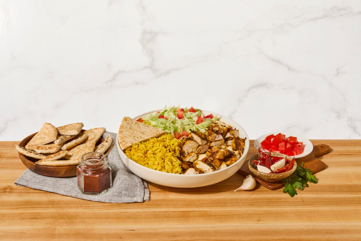 A bowl of halal chicken with yellow rice, lettuce, tomatoes, white sauce, red sauce, and pita bread.