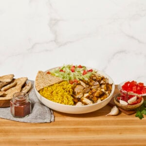 A bowl of halal chicken with yellow rice, lettuce, tomatoes, white sauce, red sauce, and pita bread.