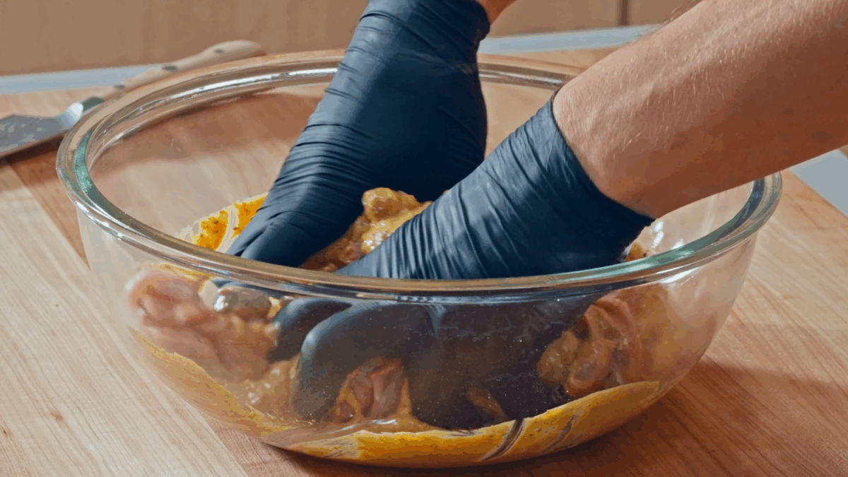 Nick DiGiovanni using gloved hands to toss raw chicken thighs in halal marinade.