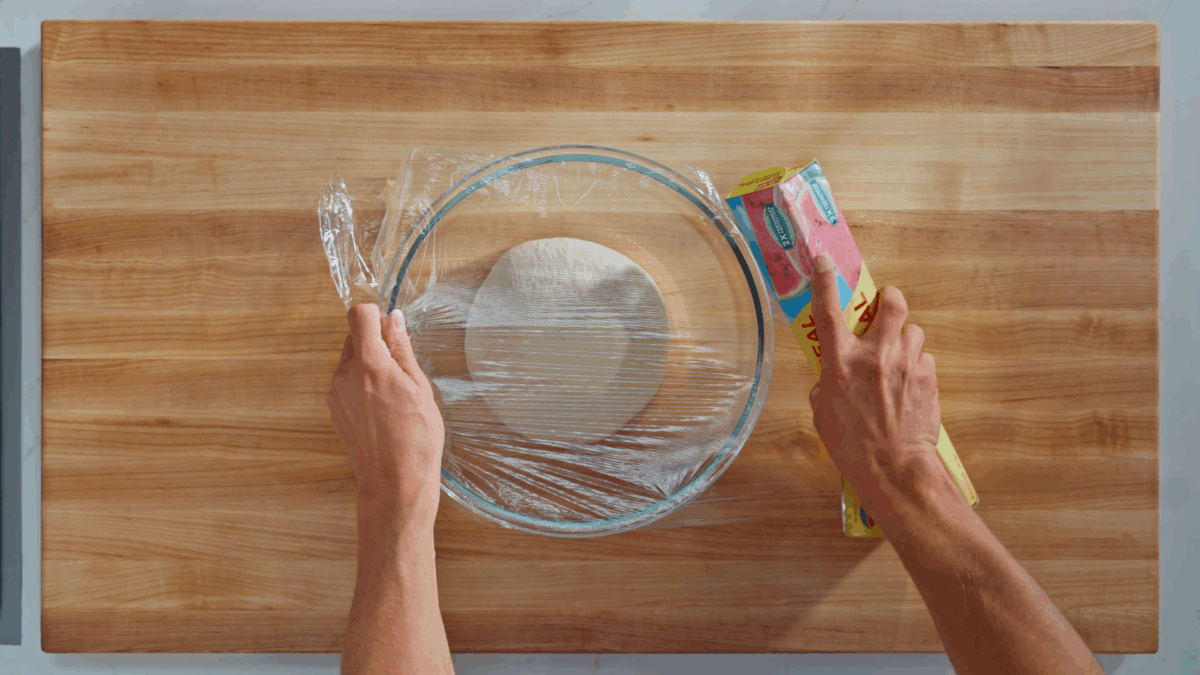 Uncooked bagel dough in a bowl being covered with plastic wrap. 
