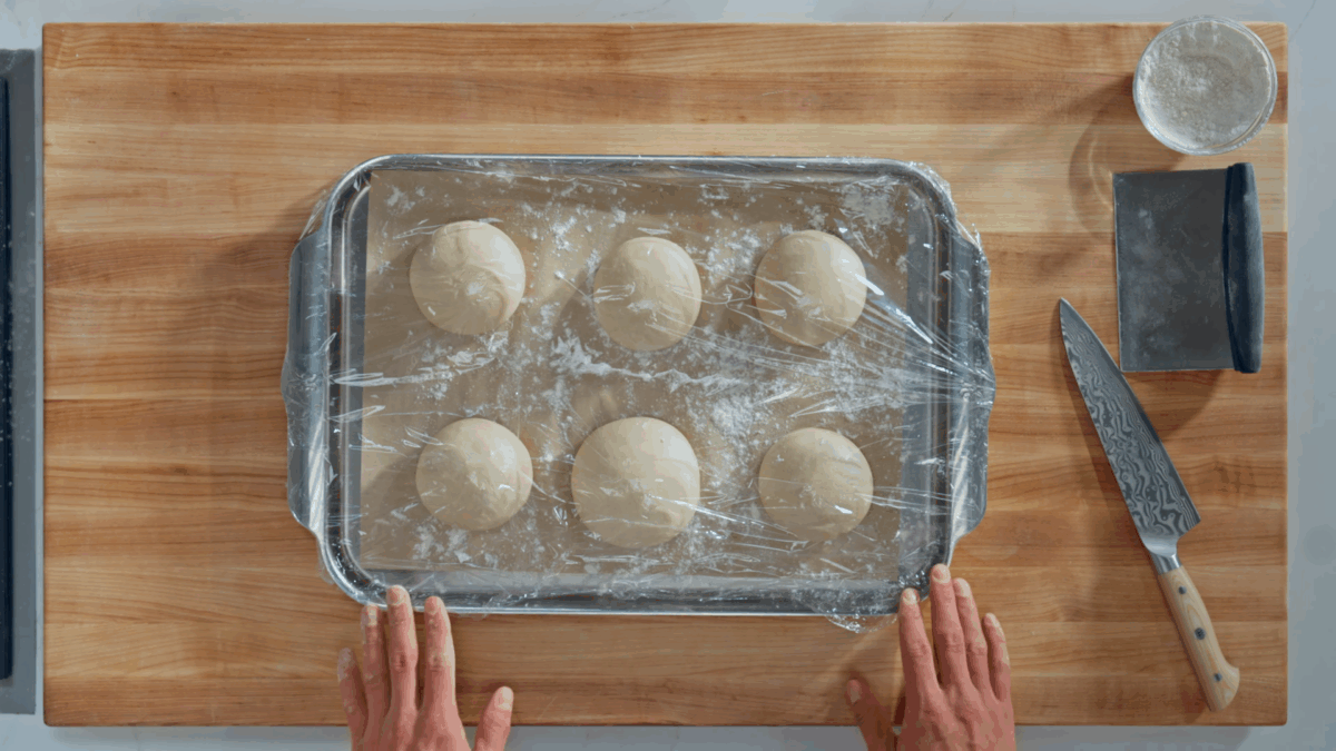 Six balls of bagel dough proofed on a parchment paper-lined baking sheet covered with plastic wrap. 