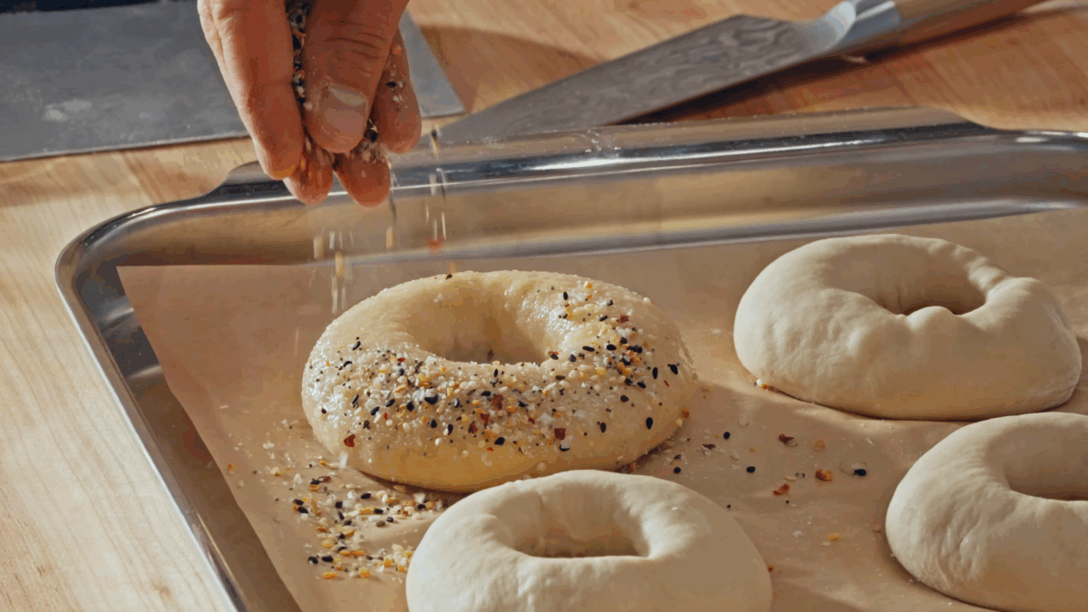 Nick DiGiovanni sprinkling everything but the bagel seasoning over wet homemade bagels on a baking sheet. 