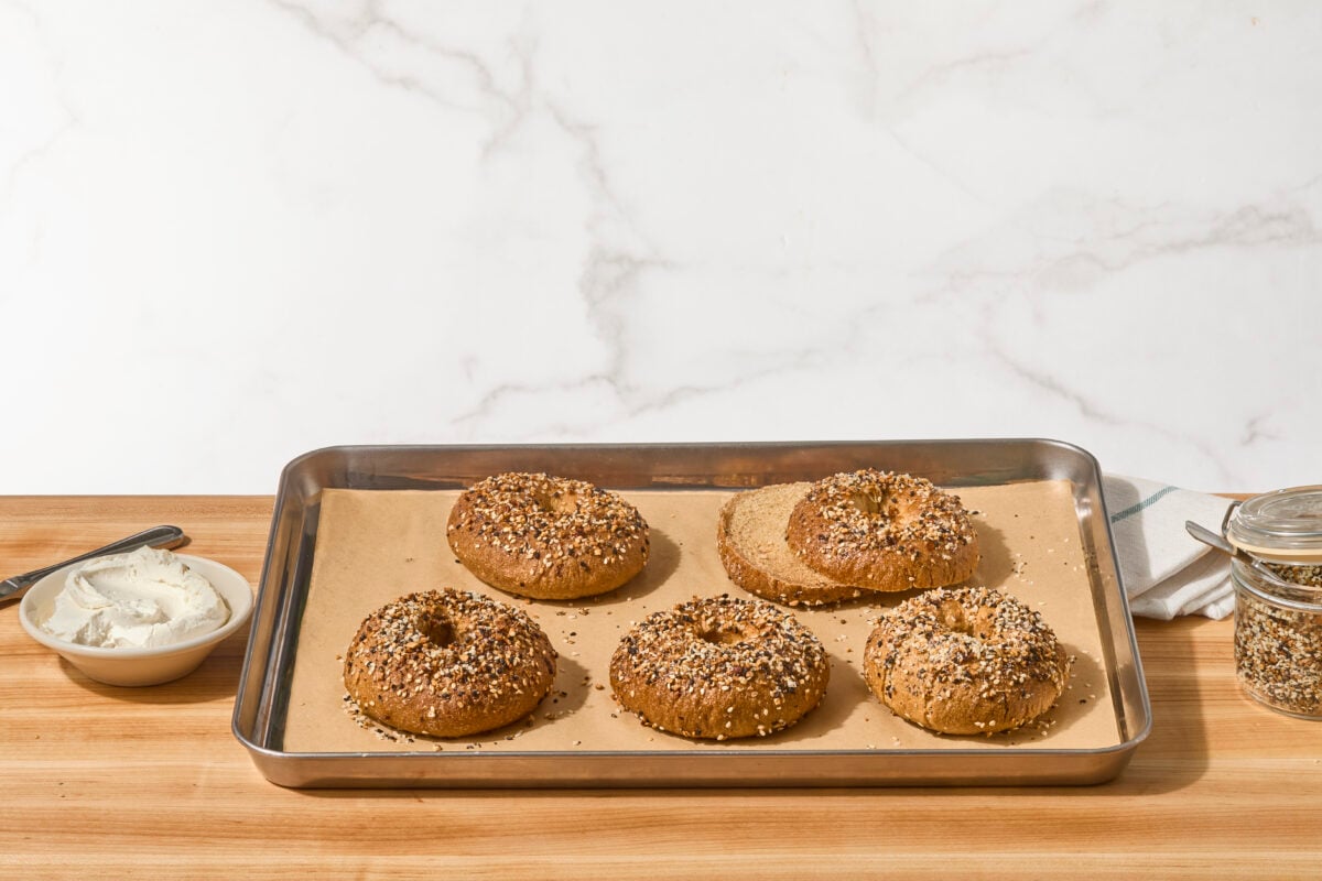 Five homemade bagels on a baking sheet, one sliced in half.