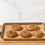 Five homemade bagels on a baking sheet, one sliced in half.