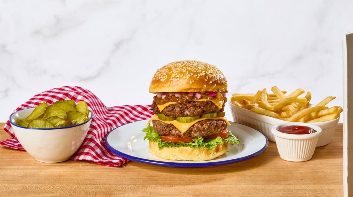 A double cheeseburger on a plate next to a bowl of pickles and fries.