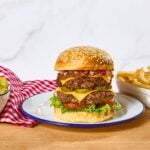 A double cheeseburger on a plate next to a bowl of pickles and fries.