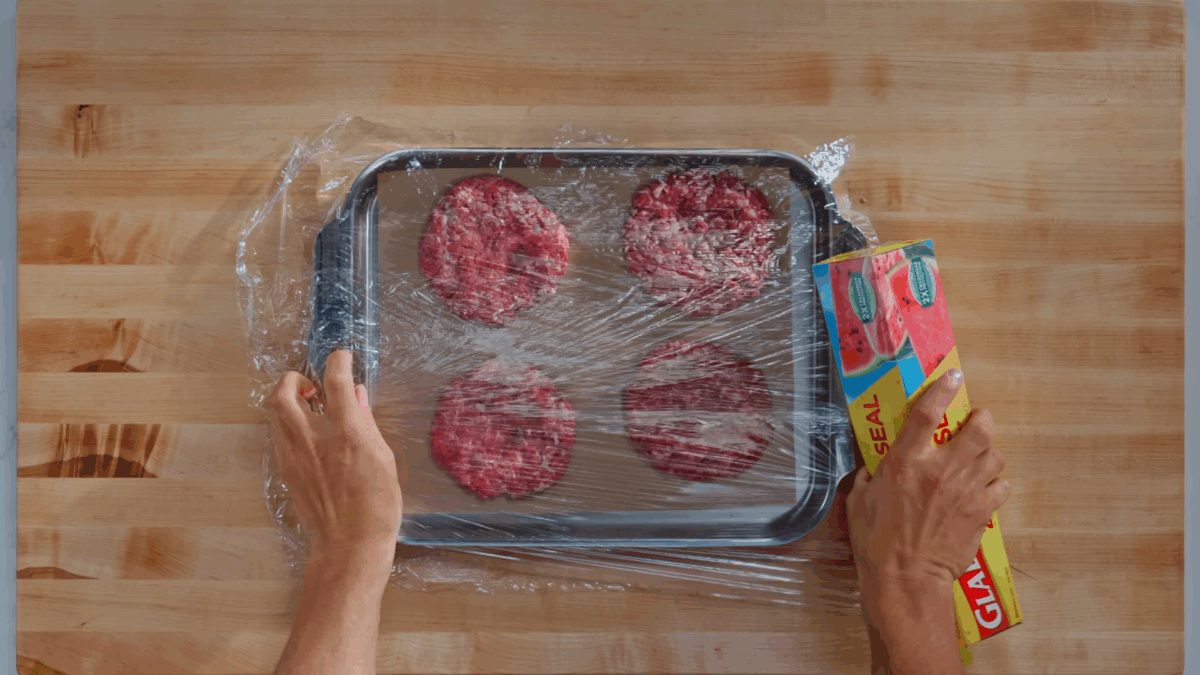 Nick DiGiovanni covering a baking sheet with four raw burger patties with plastic wrap.