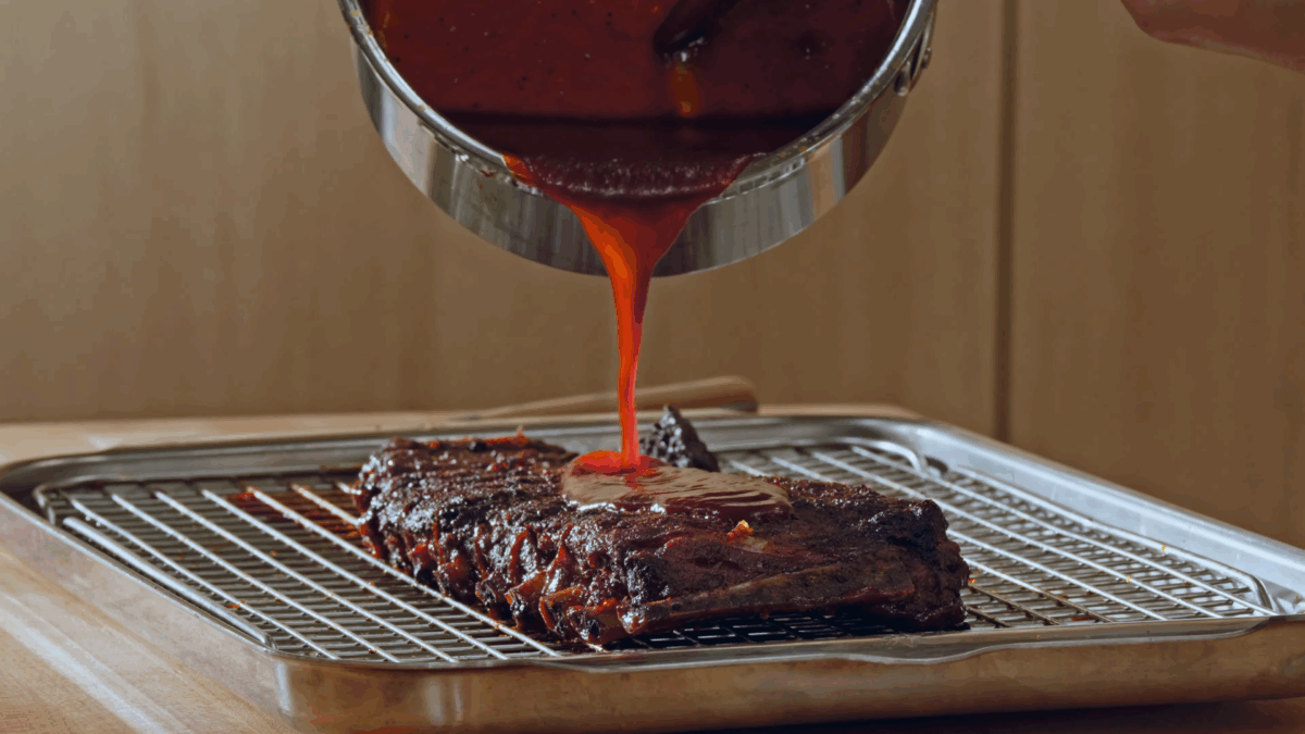 Nick DiGiovanni pouring BBQ sauce over a rack of St. Louis style pork ribs on a wire rack lined sheet tray.