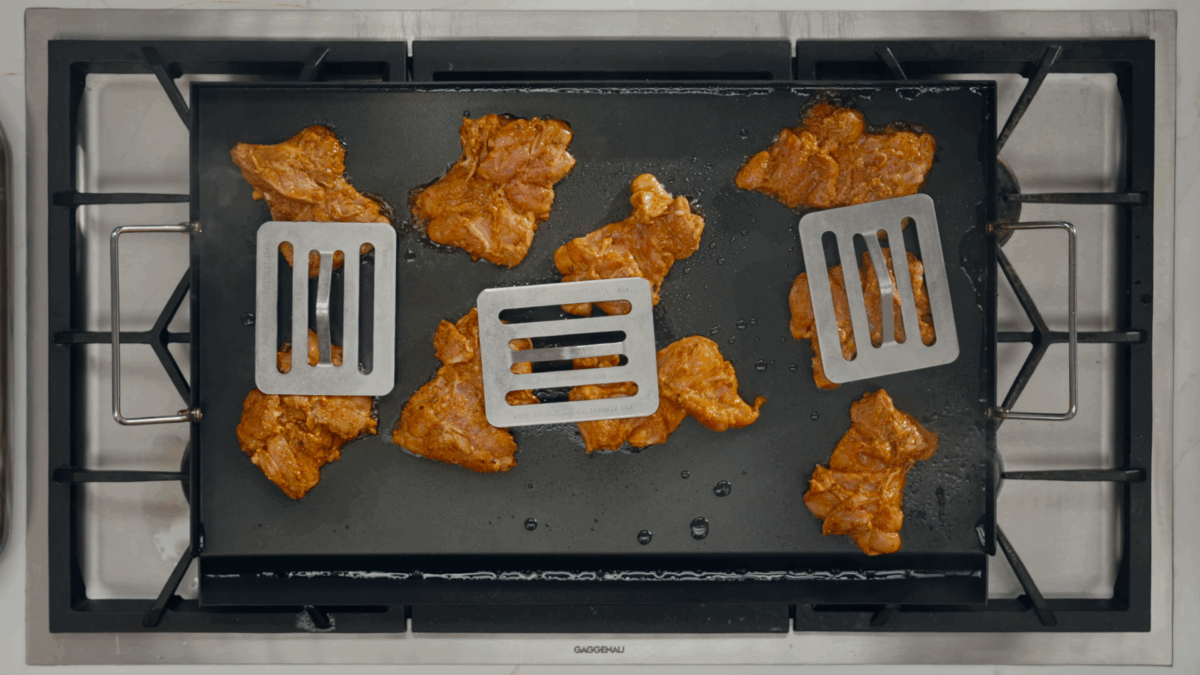 Marinated chicken cooking on a flat top pressed down by kitchen weights. 