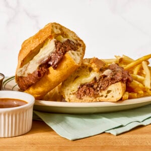Two halves of a French dip sandwich on a plate with fries, a cup of au jus next to it.