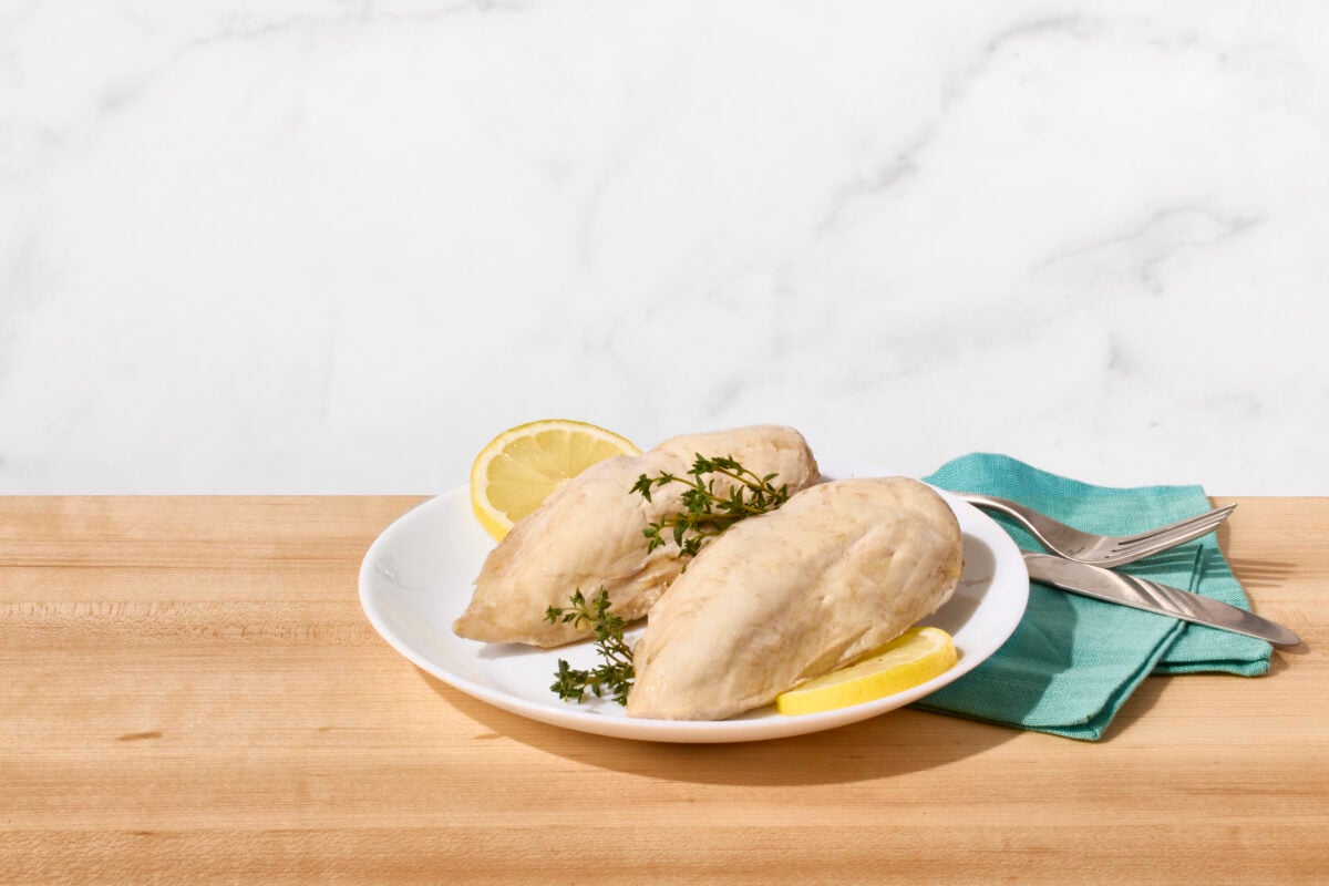 Two poached chicken breasts on a plate with fresh herbs and lemon slices.