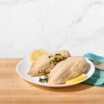 Two poached chicken breasts on a plate with fresh herbs and lemon slices.